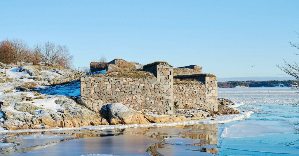 winter landscape featuring a stone structure on the snowy and rocky shore, with a partially frozen body of water in the foreground and clear blue skies above