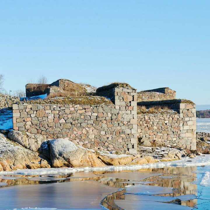 winter landscape featuring a stone structure on the snowy and rocky shore, with a partially frozen body of water in the foreground and clear blue skies above